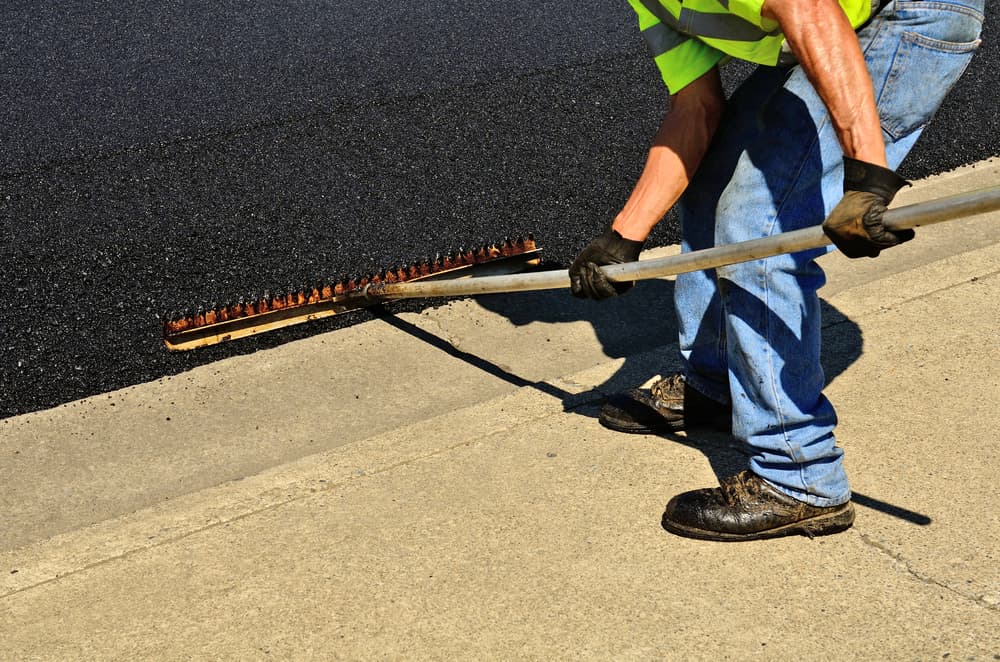 Worker using a rake to push excess asphalt off of concrete curbing on a repaving construction project